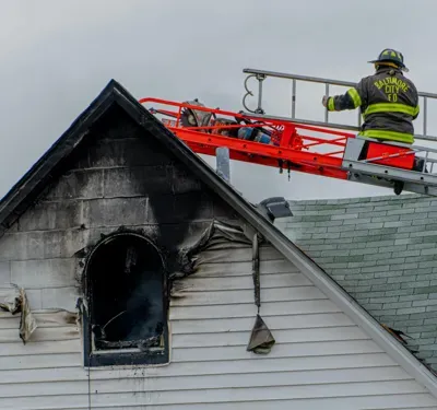 Firefighter approaches smoke and fire damaged house in crane bucket.